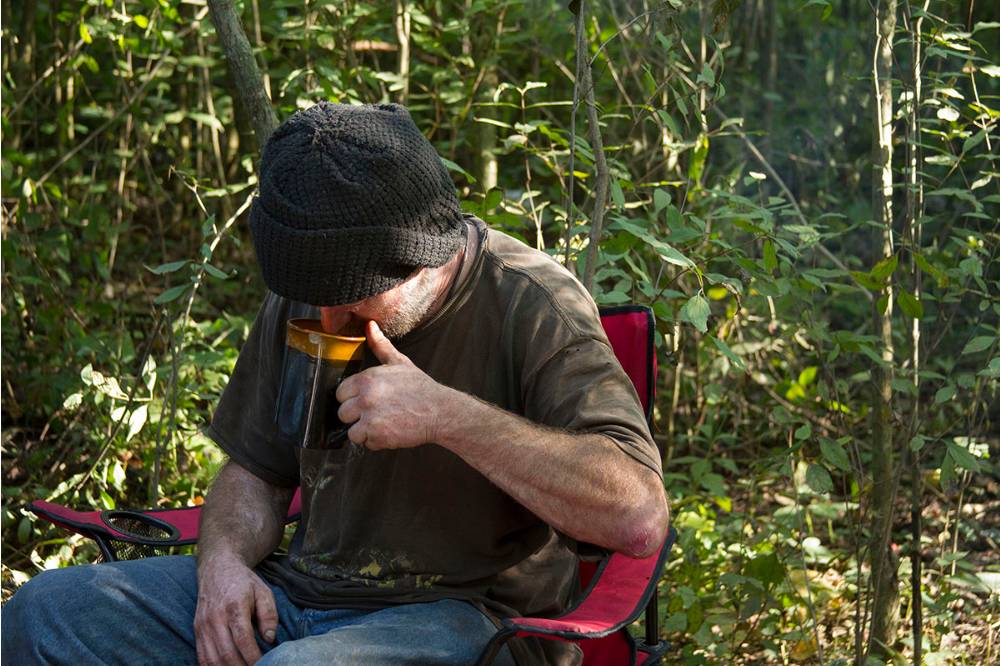 Man wearing black hat, sitting in camp chair in woods drinking from a coffee cup